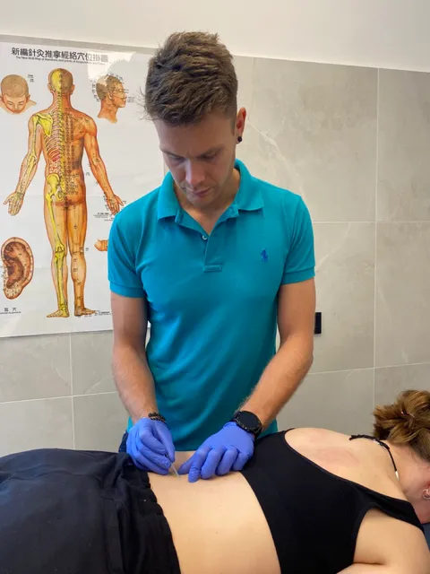 Paweł Grajewski inserting acupuncture needle with Chinese medicine chart in the background